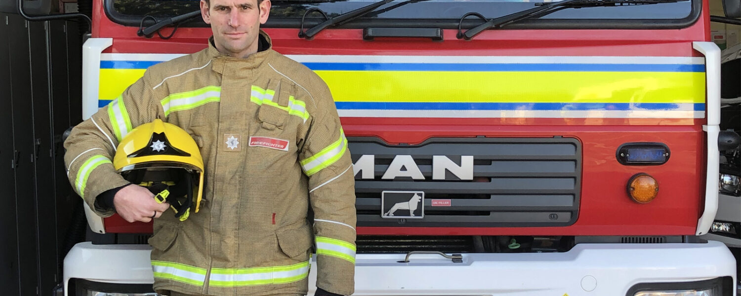 white male firefighter in full kit, holding helmet to his side. In front of fire engine