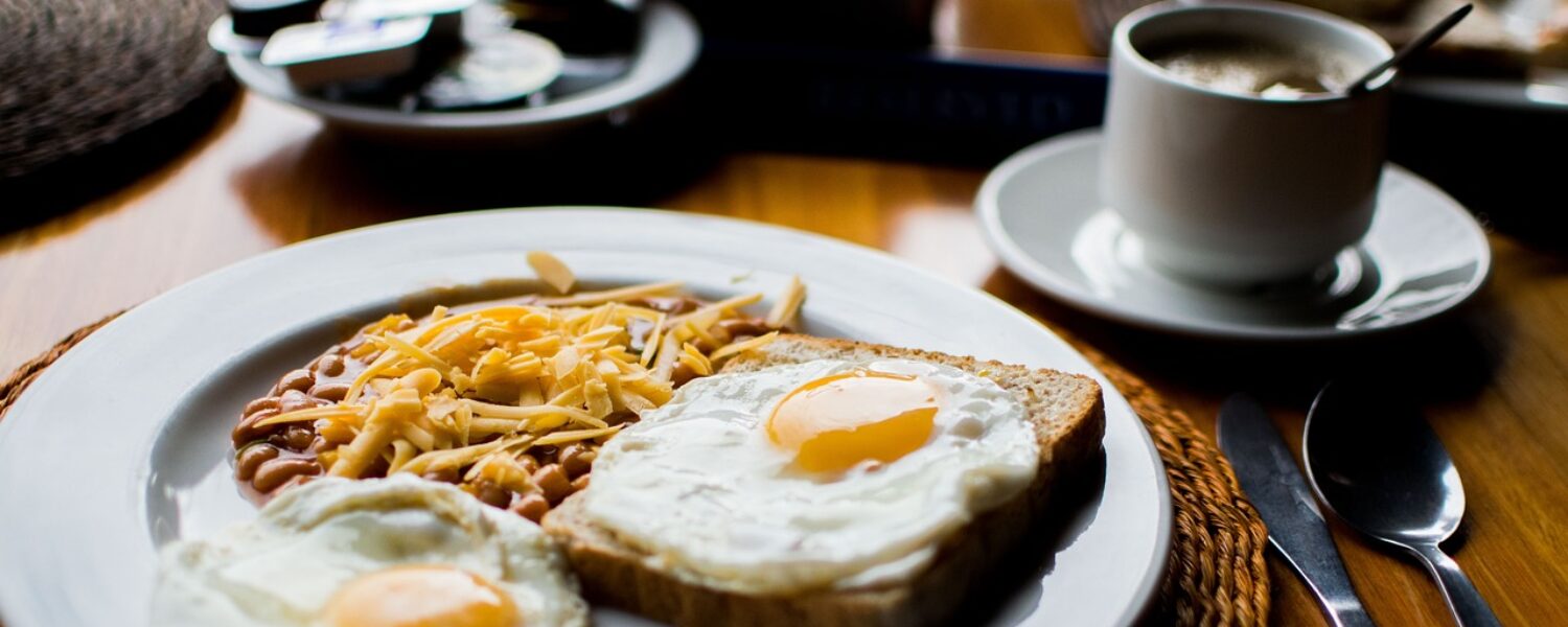 table with plate contained beans, and two fried eggs with one of them on bread. Coffee in a white cup with a saucer to the right of the plate. Small plate containing sachets of jams and spreads.