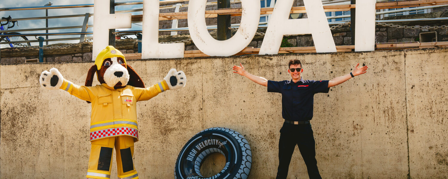 fido the firedog large dog mascot standing next to a firefighter both doing the 'float to live' position by stretching out their arms and legs. They are on a beach, with the word 'float' behind them.